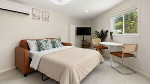 A modern bedroom with a bed, a TV, a small table, and a window with a view of greenery. - Pool and Pool House in McAllen (McAllen)