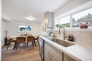A modern kitchen with a built-in sink, a dining area with a table and chairs, and a window with a view of trees and a building. - Family-Friendly 4BR Retreat, Near UW & Parks (Seattle)