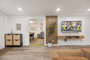A modern living room with a flat-screen TV mounted on the wall, a potted plant, and a dining area in the background. - Family-Friendly 4BR Retreat, Near UW & Parks (Seattle)