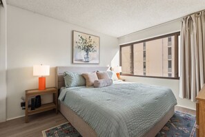 A bedroom with a bed, two bedside tables with lamps, a painting on the wall, and a window with curtains. - Waikiki Hideaway · Canal Views•Pool•Parking•W/D (Honolulu)
