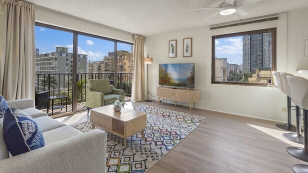 A modern living room with a sofa, armchair, coffee table, TV, and a balcony with a city view. - Waikiki Hideaway · Canal Views•Pool•Parking•W/D (Honolulu)