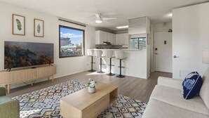 A modern apartment with a living room, kitchen, and dining area. There is a large window, a ceiling fan, and a TV mounted on the wall. - Waikiki Hideaway · Canal Views•Pool•Parking•W/D (Honolulu)