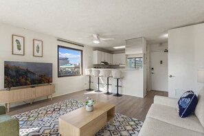 A modern apartment with a living room, kitchen, and dining area. There is a large window, a ceiling fan, and a TV mounted on the wall. - Waikiki Hideaway · Canal Views•Pool•Parking•W/D (Honolulu)