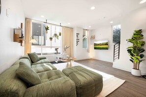 A modern living room with a sectional sofa, a wall-mounted TV, a bike, and a large window with curtains. - Modern 3BR City View Home w/ Near Green Lake & UW (Seattle)