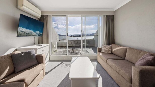 A hotel room with a large sliding glass door, a flat-screen TV, a sofa, a coffee table, and a view of a dining area outside. - Horizons 302 - Centrally located in town with pool access, tennis courts and undercover car parking (Jindabyne)