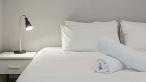 A neatly made bed with white linens, a folded towel, and a bedside lamp on a white nightstand. - Horizons 302 - Centrally located in town with pool access, tennis courts and undercover car parking (Jindabyne)