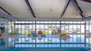 A large indoor swimming pool with clear glass walls, multiple lounge chairs, and a ceiling with a grid pattern. - Horizons 302 - Centrally located in town with pool access, tennis courts and undercover car parking (Jindabyne)