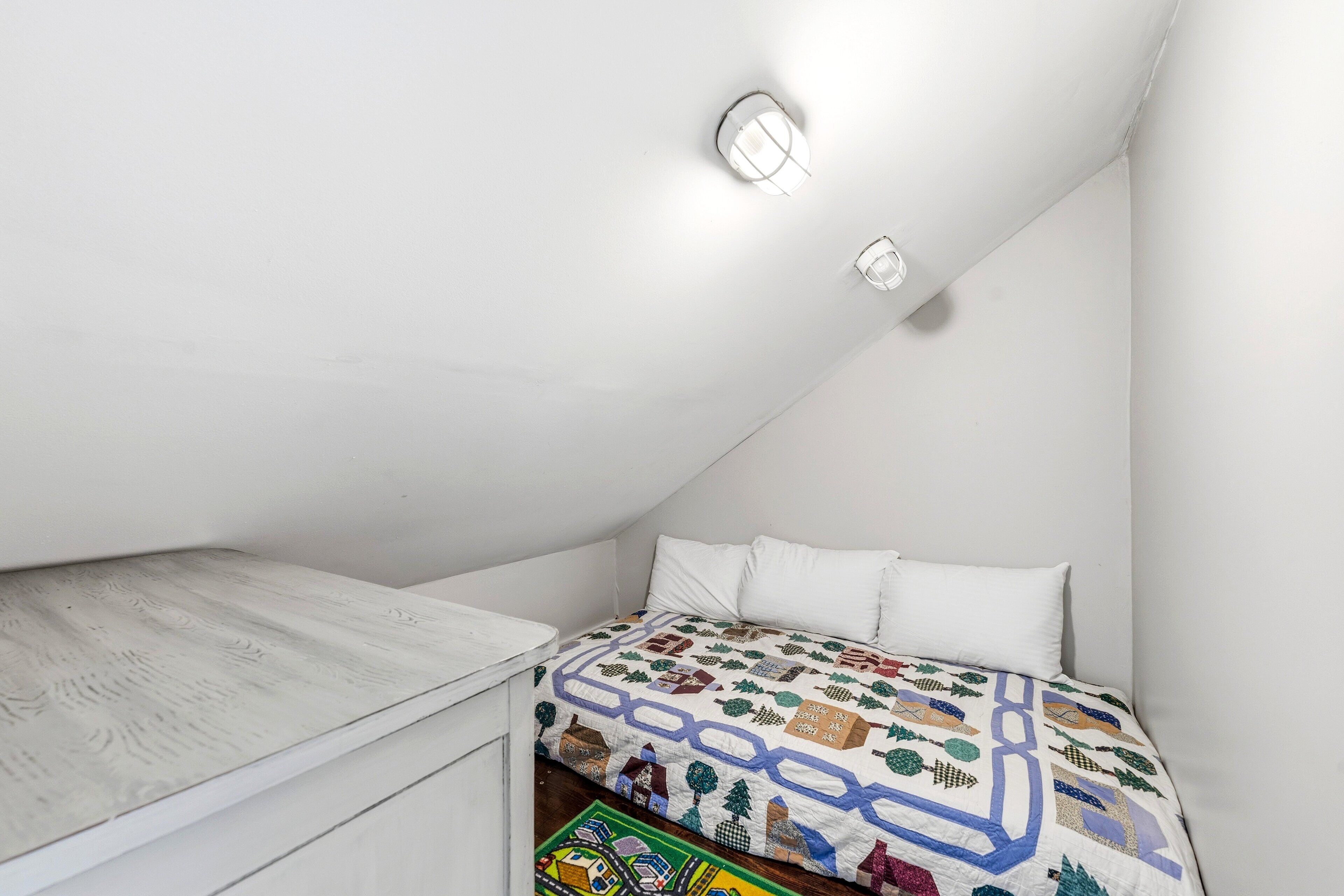 A bedroom with a sloped ceiling, a bed with a colorful quilt, white walls, and two wall-mounted lights.
