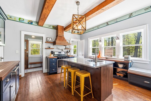A modern kitchen with wooden beams, a large island with stools, a built-in stove, and a window with a view of trees.