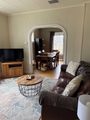 A living room with a TV, wooden entertainment unit, round coffee table, leather sofa, dining table with chairs, and a view through an archway.