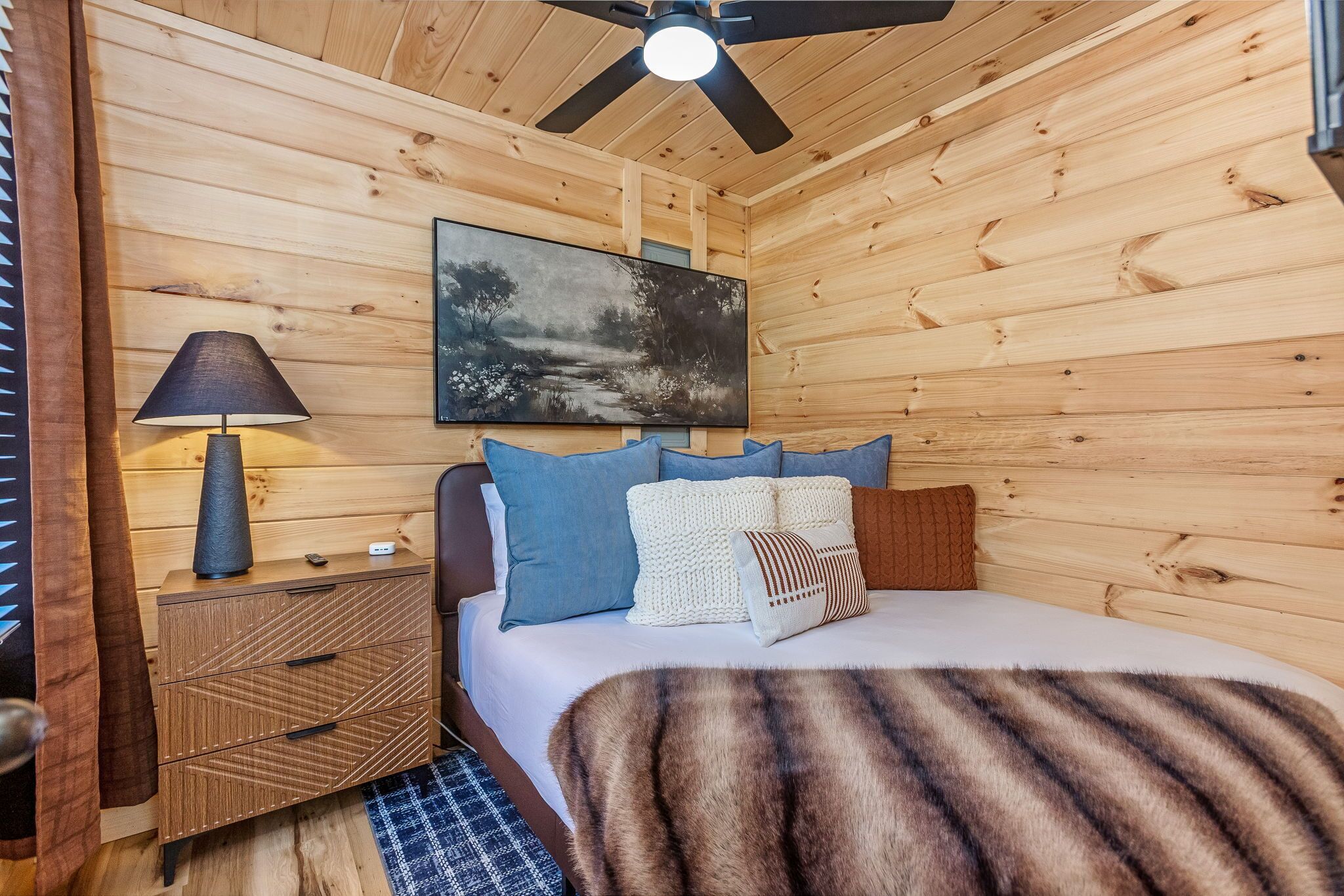 Ein Zimmer einer Holzhütte mit einem Bett, einem Nachttisch, einer Lampe und einem Deckenventilator.