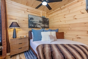 A wooden cabin room with a bed, a nightstand, a lamp, and a ceiling fan.
