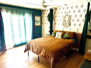 Ein Schlafzimmer mit Bett, Vorhängen, Deckenventilator, einer Wand mit strukturiertem Wandpapier und einem gerahmten Bild.