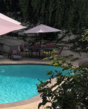 A swimming pool with a wooden deck, surrounded by chairs and tables under umbrellas, with a wall covered in ivy.
