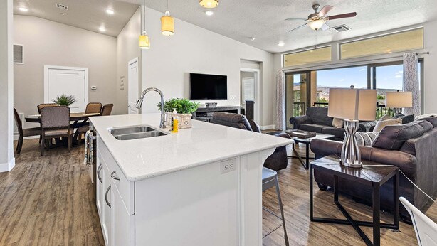 A modern kitchen with a central island, dining area with chairs, a sofa, and a TV in the background. - Estancia Resort Unit #404 (Saint George)
