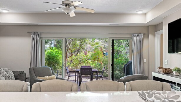 Ein modernes Wohnzimmer mit Esstisch, Stühlen, Deckenventilator und Blick auf Grünflächen durch große Schiebetüren.