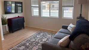 A living room with a flat-screen TV, a dark-colored sofa, a patterned rug, and a window with blinds. - Your Indio retreat awaits you (Indio)