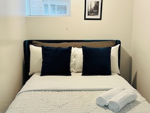 A bed with a white and dark blue headboard, two pillows, a rolled white towel, and a framed picture on the wall.