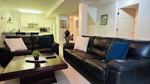 A living room with a dark brown leather sofa, a coffee table with a potted plant, and a kitchen in the background.