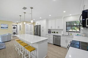 Eine moderne Küche mit weißen Schränken, einer zentralen Kochinsel mit Hockern, einem eingebauten Backofen und einem Edelstahlkühlschrank.