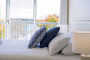 Een hotelkamer met een bed, kussens, een lamp en uitzicht op bomen door grote ramen.