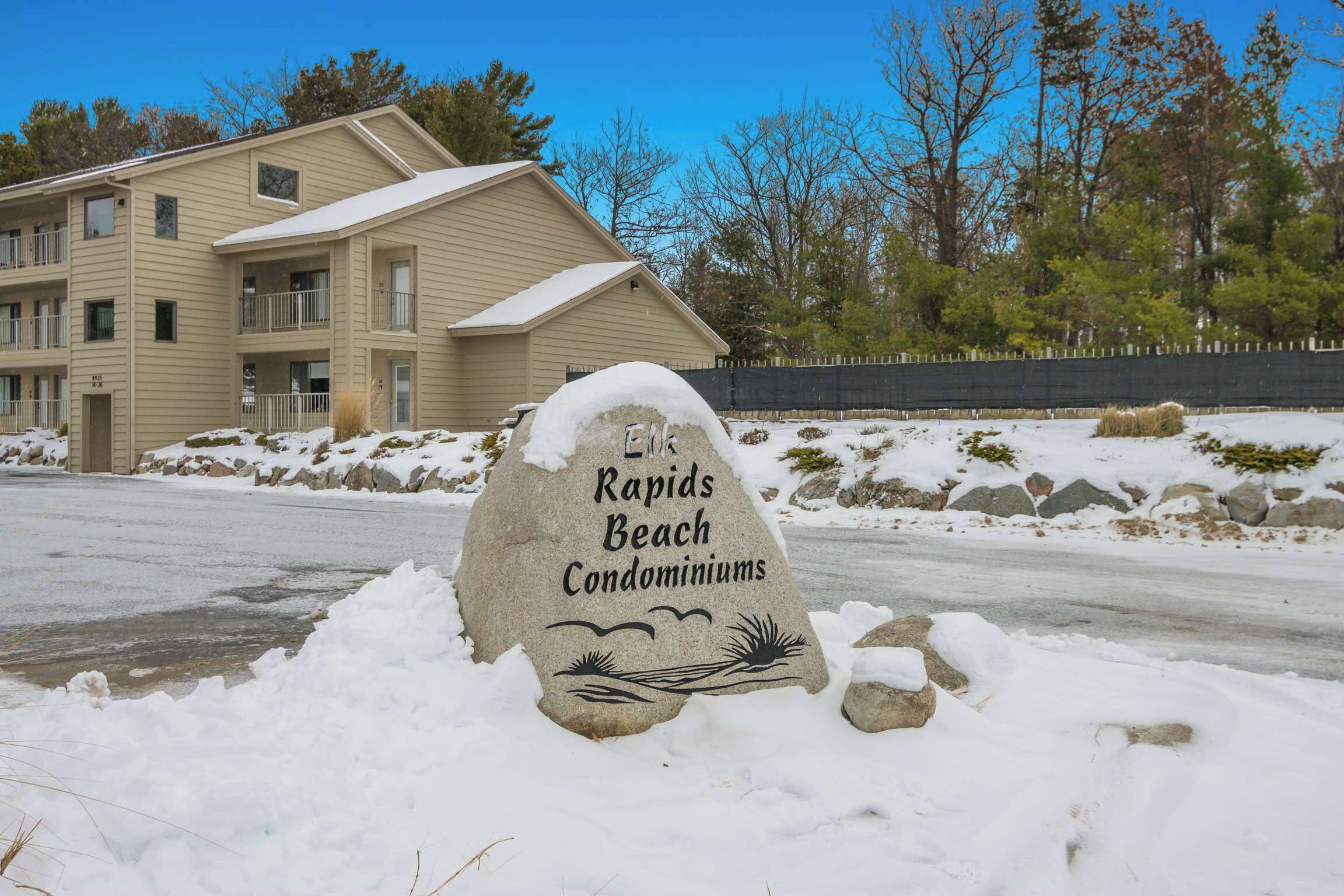 Un senyal amb mantell de neu anuncia Rapids Beach Condominiums, amb un edifici de diversos pisos al fons.