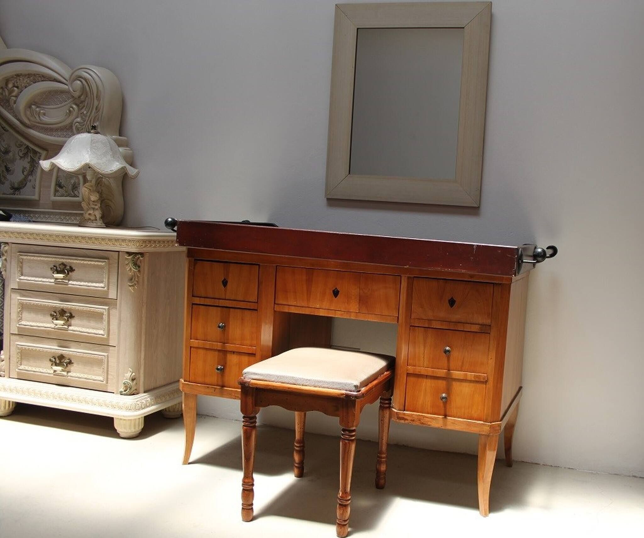 A wooden desk with a cushioned chair, a dresser with drawers, and a framed mirror on the wall.
