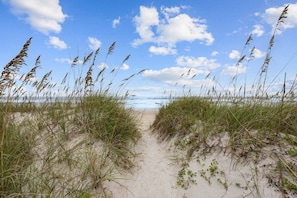 Ein sandiger Pfad führt zum Strand, begleitet von hohem Gras an beiden Seiten.