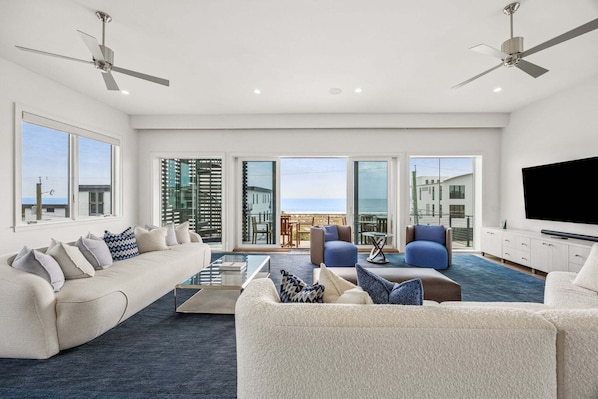 A spacious living room with a large sofa, a coffee table, and a flat-screen TV. There are ceiling fans, large windows, and a balcony with a view.