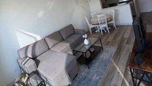En moderne stue med en grå sofa, et sofabord og et fjernsyn. Der er et spiseområde med et hvidt bord og stole. Gulvet er i træ, og der er en loftlampe.