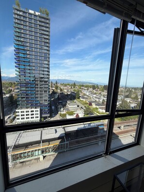 A high-rise building with multiple balconies, a clear sky, and a distant view of a town with houses and a train track. -    Live in a Safe Neighborhood, Steps to SkyTrain & Major Bus Routes (Vancouver)