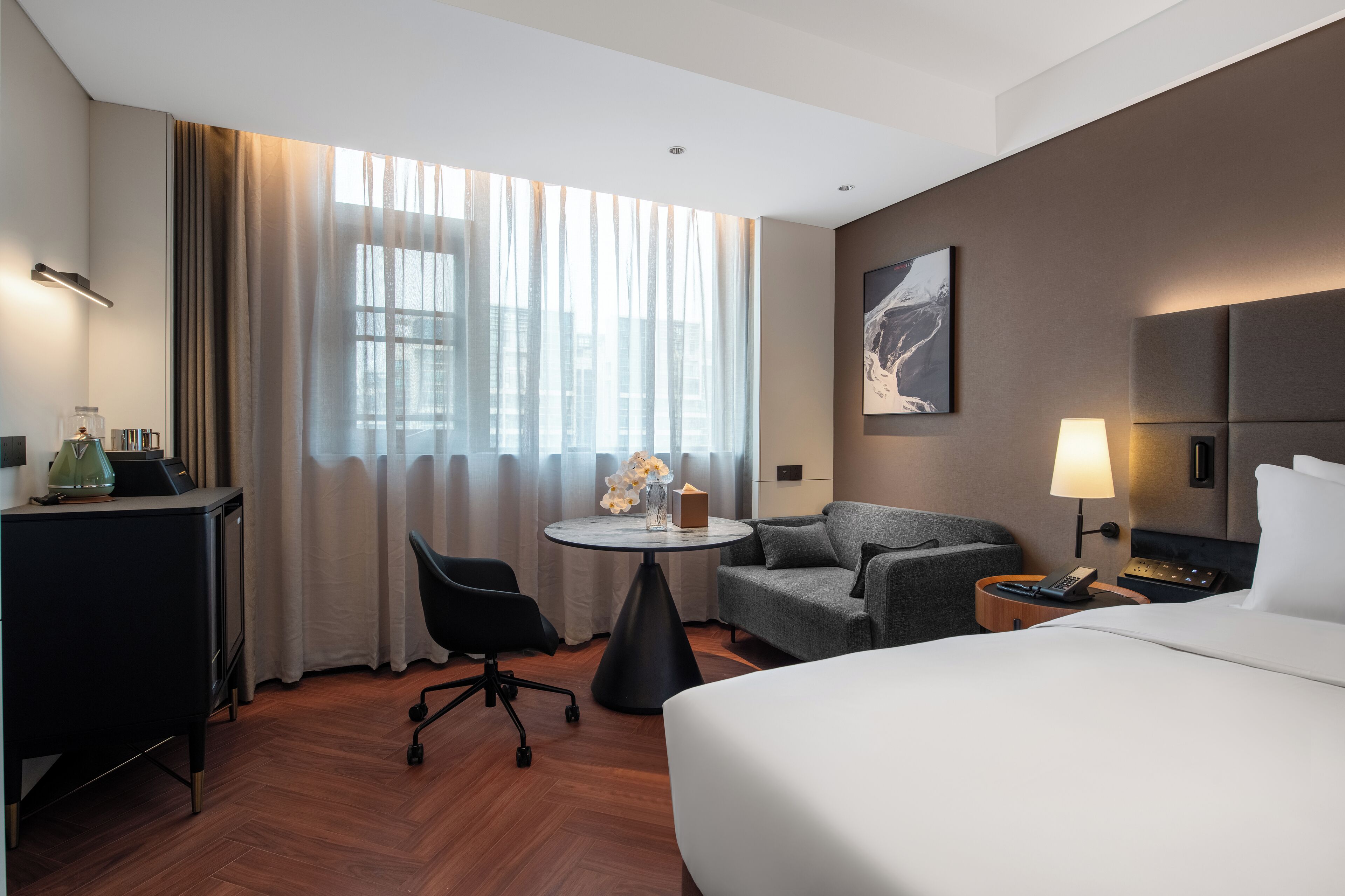 A hotel room with a bed, a desk, a chair, a small table with flowers, a lamp, and a painting on the wall.