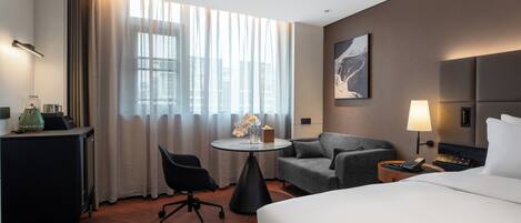 A hotel room with a bed, a desk, a chair, a small table with flowers, a lamp, and a painting on the wall.