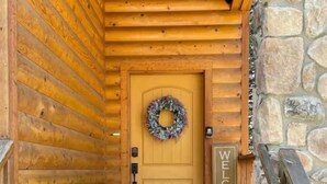En entré till en stuga byggd av trästockar, med en stenmur, en trädörr prydd med en krans och en skylt med texten ”VÄLKOMMEN”.