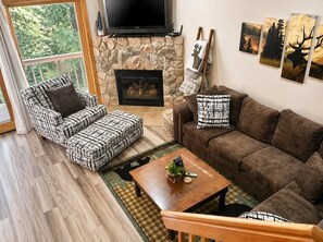 Ein Wohnzimmer mit Kamin aus Stein, einem darüber hängenden Fernseher, einem braunen Sofa, gemusterten Sessel und einem Couchtisch mit Pflanze.