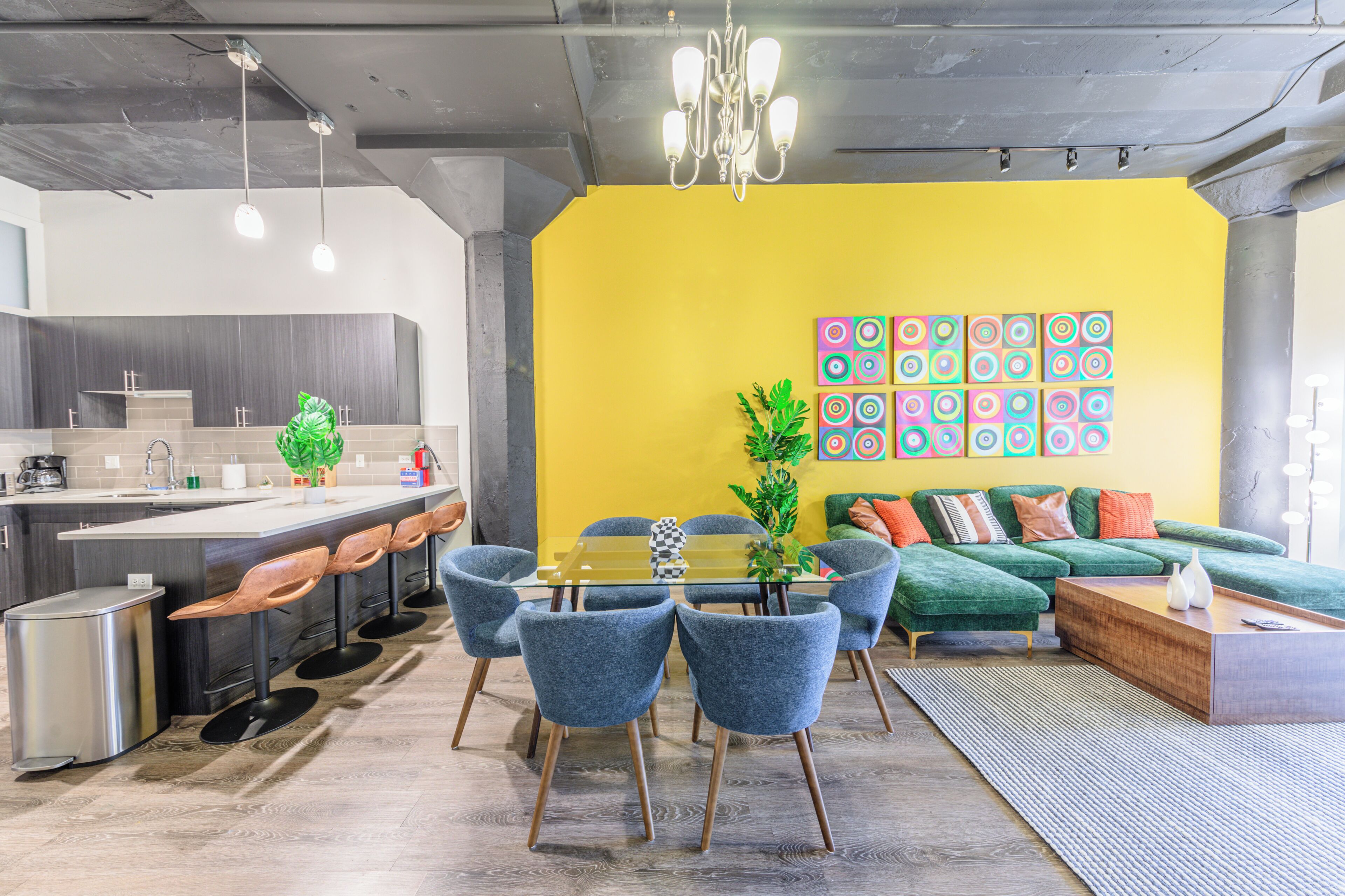 A modern kitchen with a bar area, a dining table with chairs, a sofa, and a colorful wall art piece.