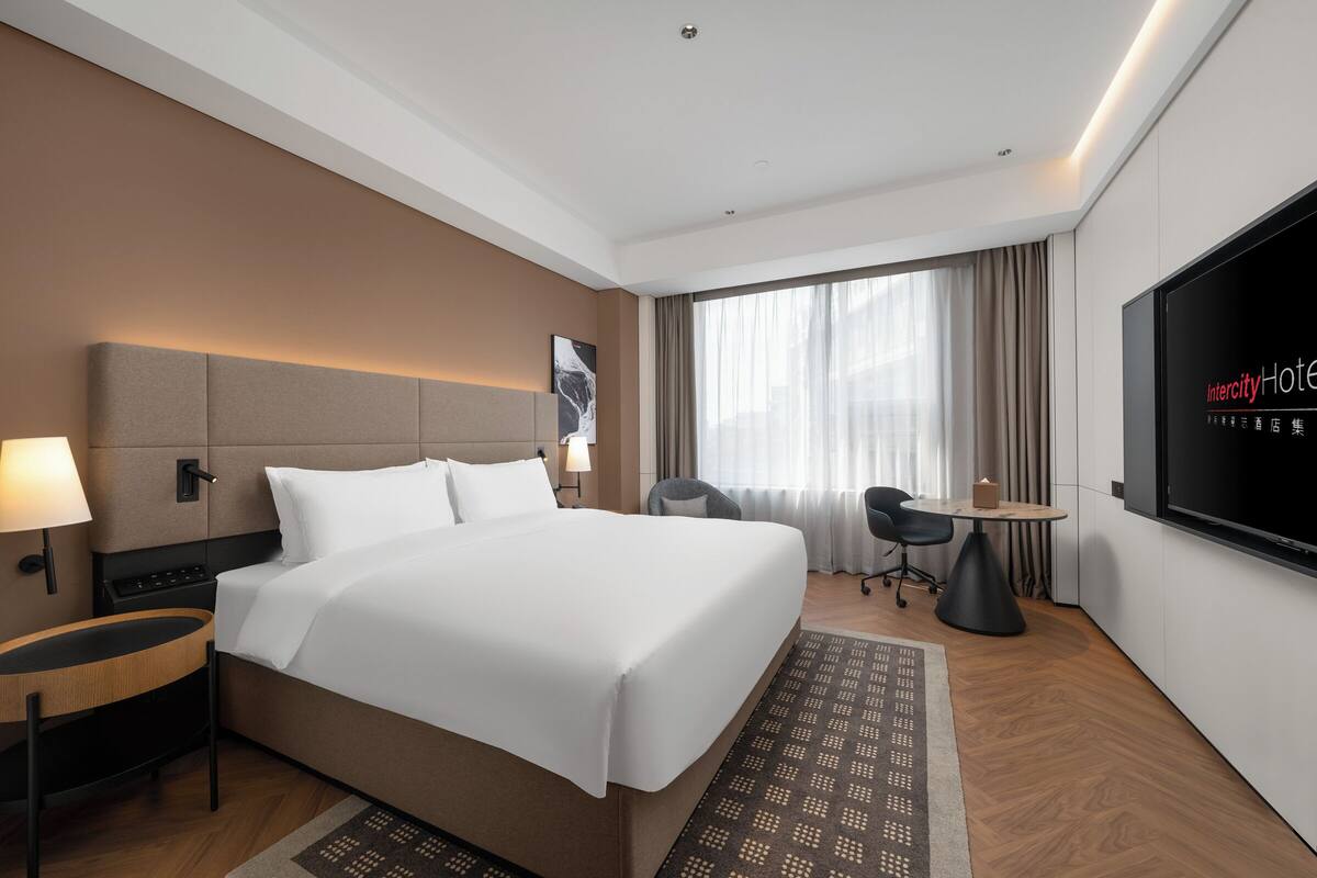 Ein Hotelzimmer mit Bett, Nachttisch, Sessel, Schreibtisch, Fernseher und Vorhängen.