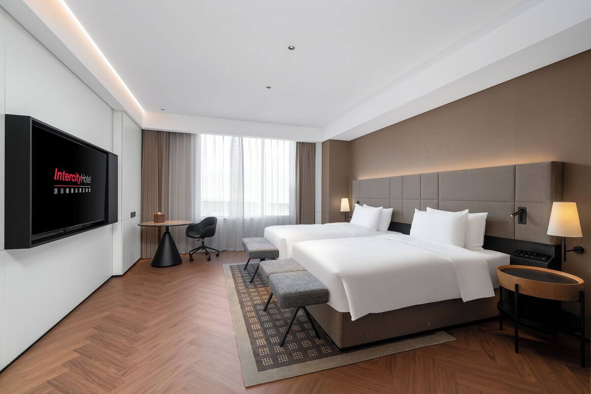 Ein Hotelzimmer mit einem großen Bett, einem an der Wand montierten Fernseher, einem Schreibtisch und einem Fenster mit Vorhängen.