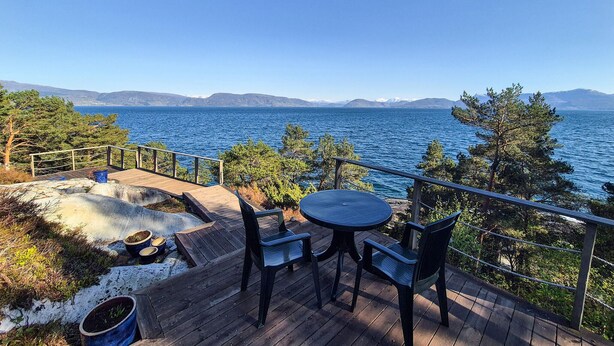 Una terraza de madera con mesa y sillas que da a un cuerpo de agua y montañas.