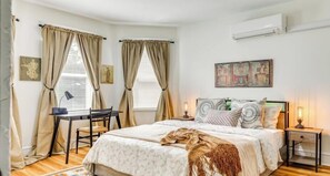 A bedroom with a bed, a desk, a chair, two windows with curtains, a wall-mounted air conditioner, and a painting. - Sports lovers, foodies, families, and college visitors will feel right at home in this top‑floor 4‑bed, 2‑bath triple‑decker with balcony, fully equipped kitchen, park views, and easy access to Boston. (Boston)