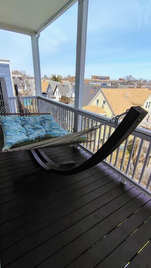 A balcony with a hammock, a cushioned seat, and a view of surrounding buildings. - Sports lovers, foodies, families, and college visitors will feel right at home in this top‑floor 4‑bed, 2‑bath triple‑decker with balcony, fully equipped kitchen, park views, and easy access to Boston. (Boston)