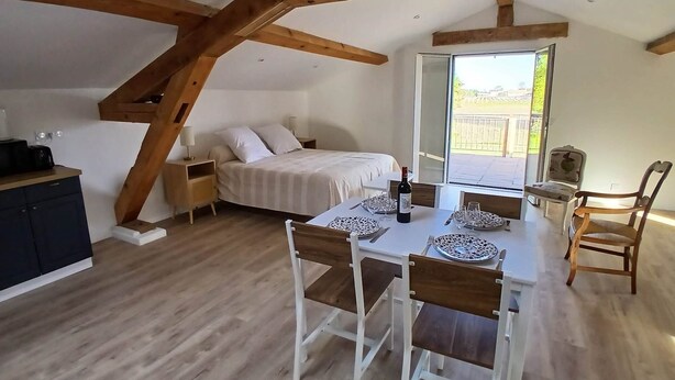 A room with a bed, a dining table with chairs, a kitchen area, and a large window.
