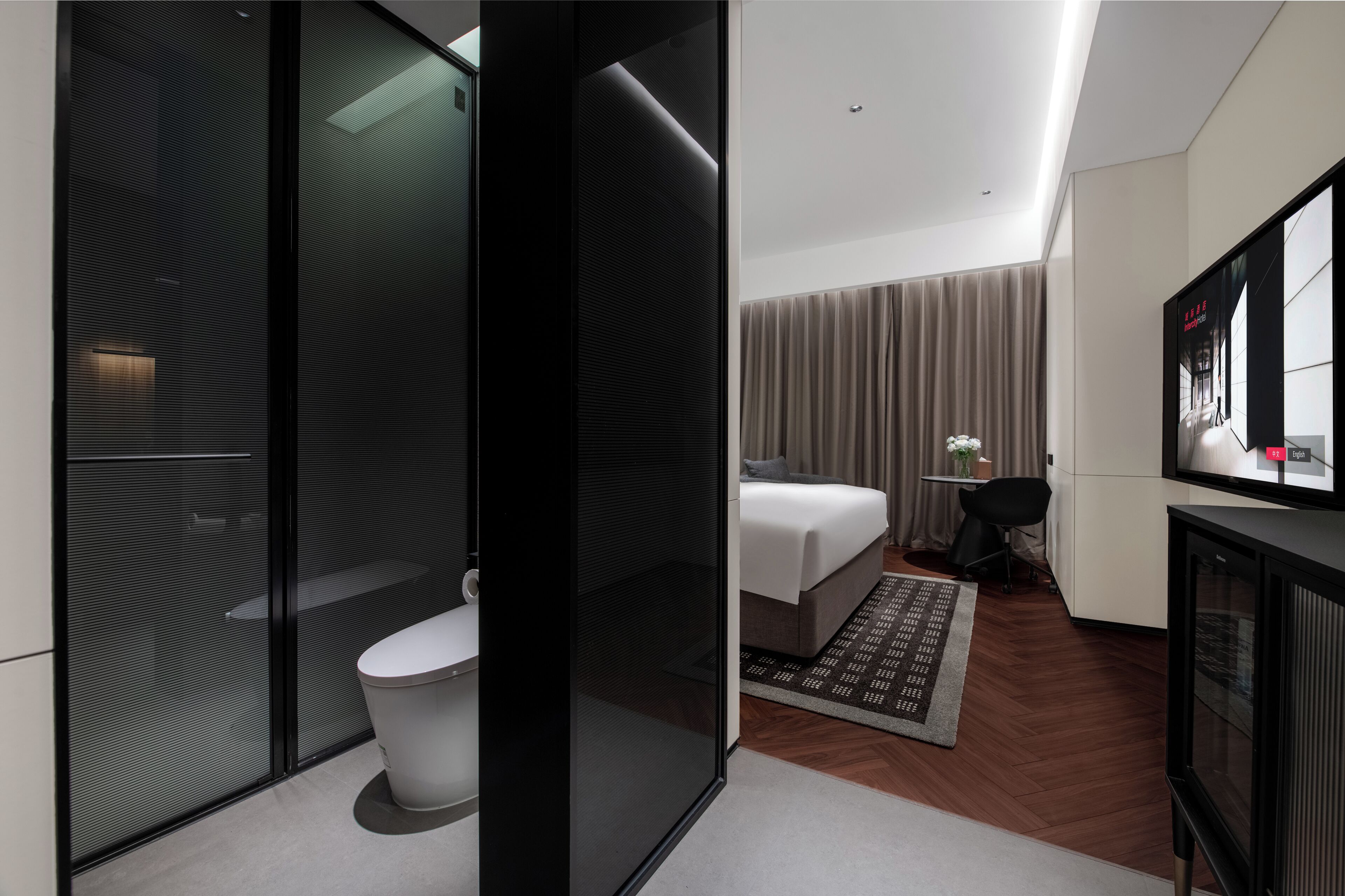 A modern hotel room with a glass partition, a white toilet, a bed, a TV, and a small table with a plant.