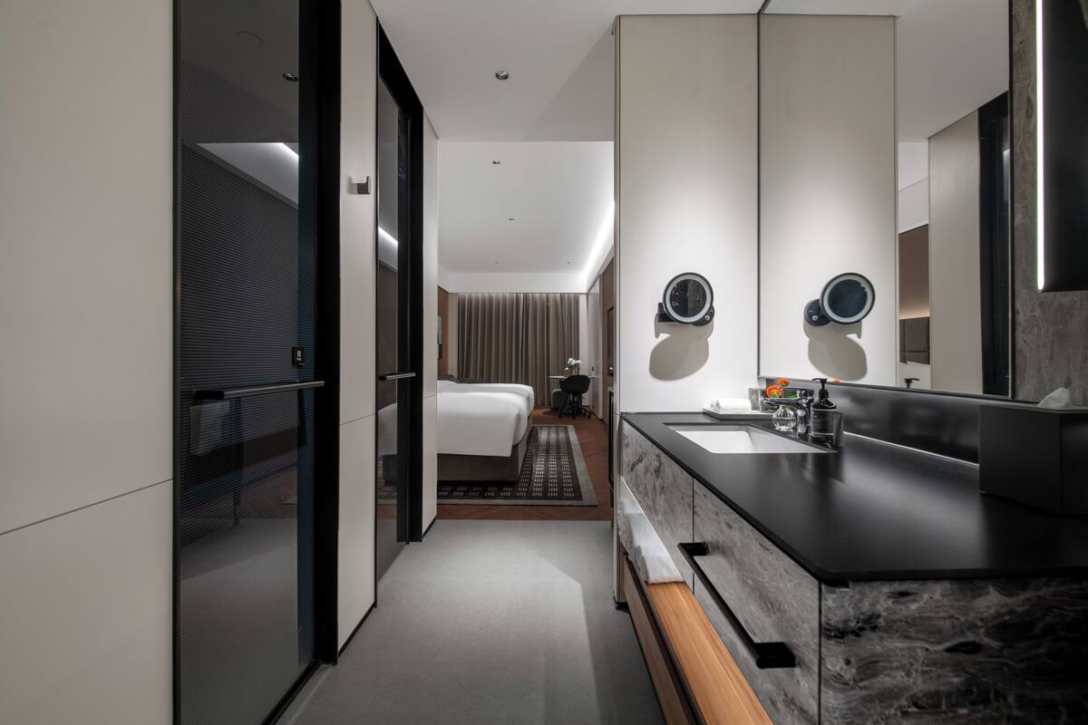 Una habitación de hotel moderna con un espejo grande, una cama, un baño con lavamanos y espejo, y una mesita con una silla.