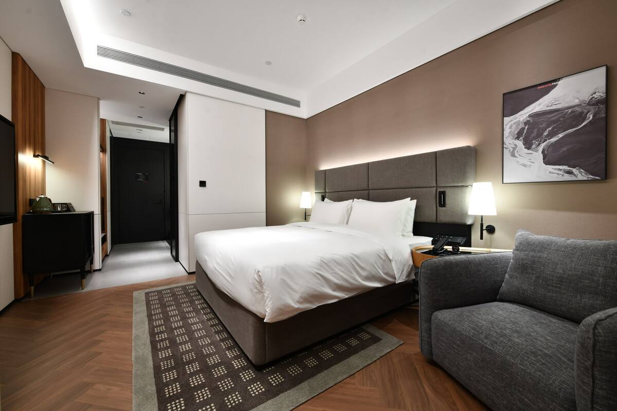 Habitación de hotel moderna con una cama grande, un sofá gris, un televisor montado en la pared y un cuadro.