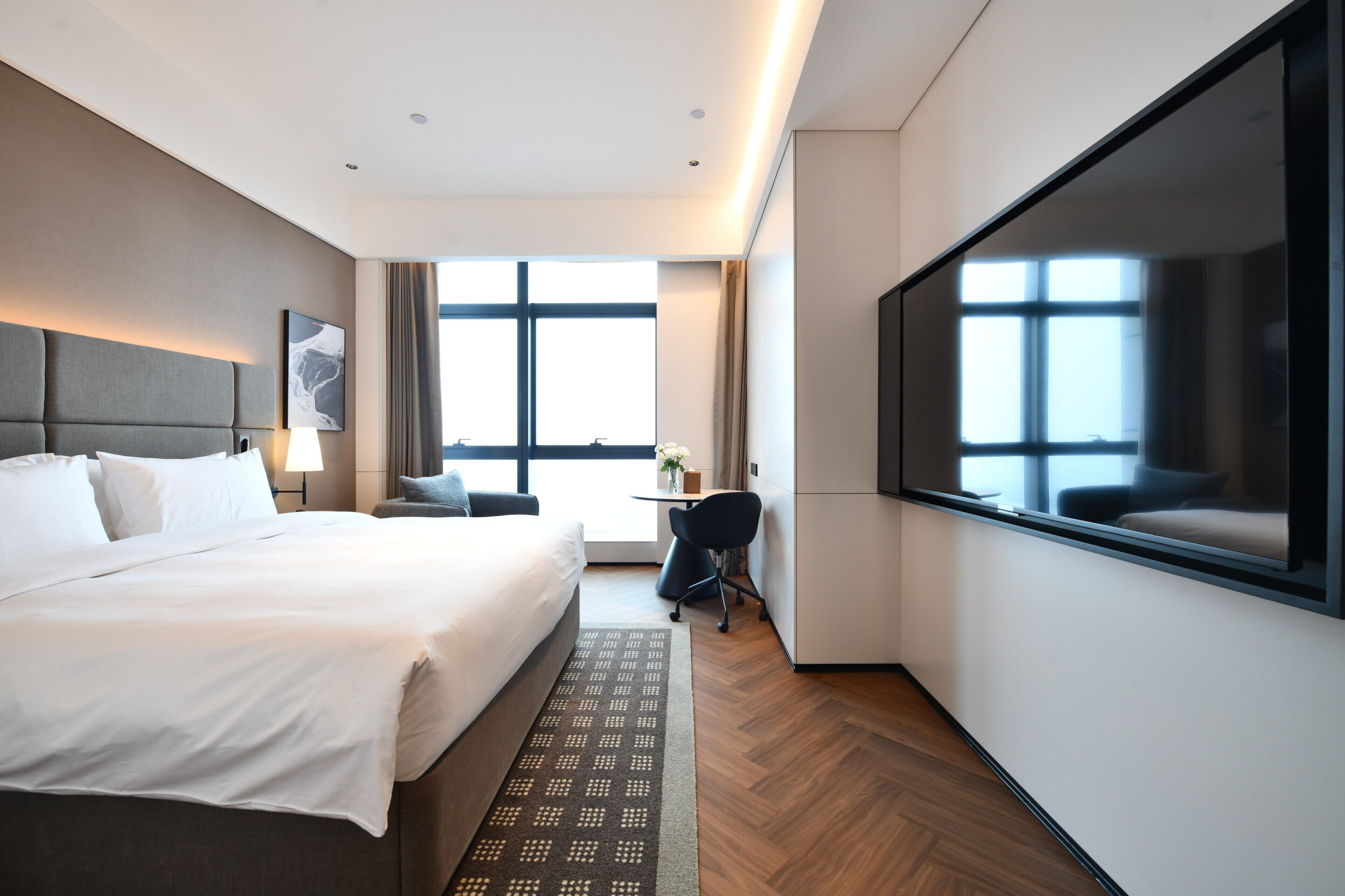 A modern hotel room with a large bed, a desk, a chair, and a large window.