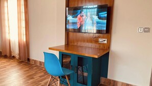 A room with a wooden floor, a blue chair, a small wooden table, a television mounted on the wall, and a power outlet. - Aronnonibash Eco Resort (Kamalganj)