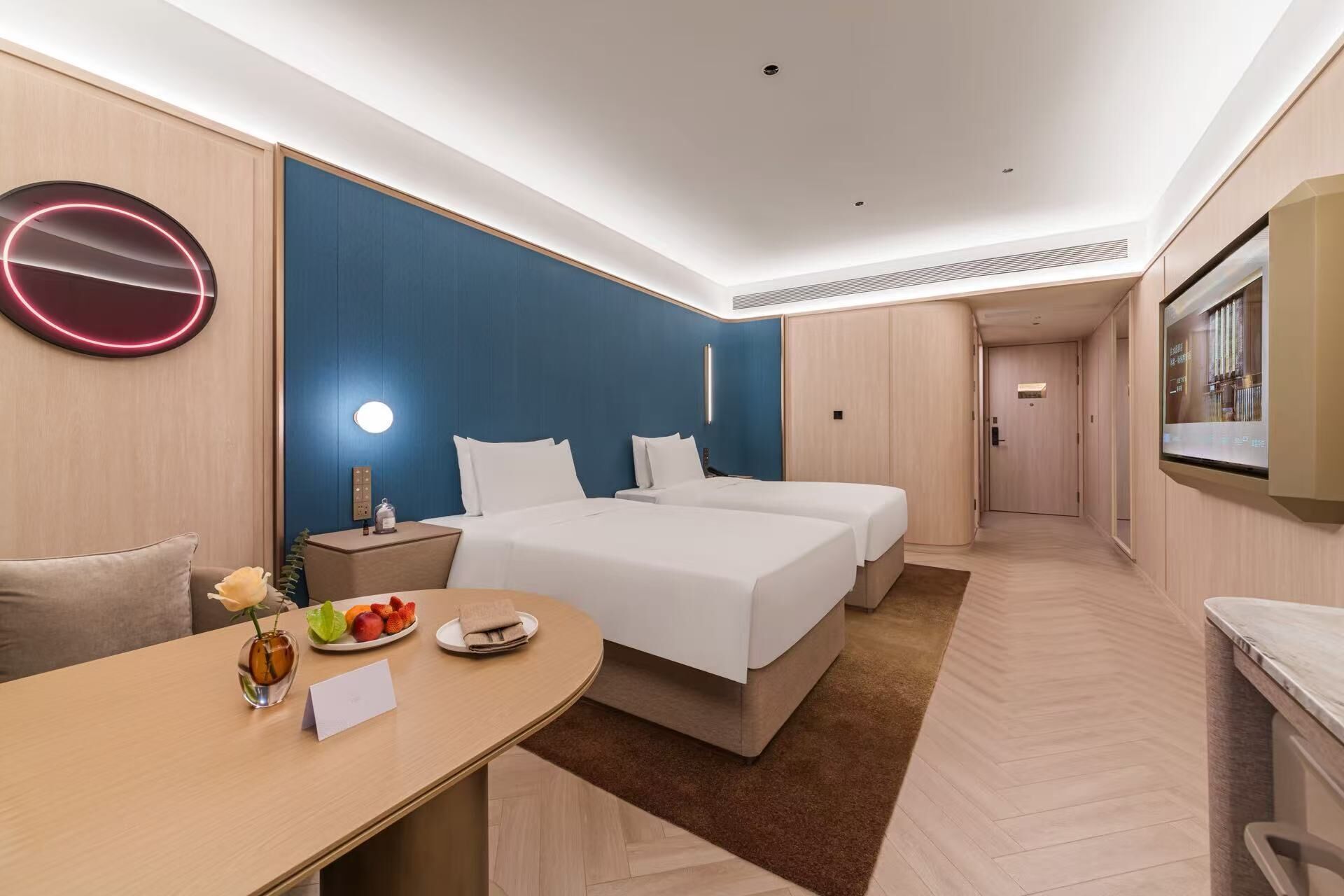 A hotel room with two beds, a desk with a vase and fruit, a round mirror, and a TV.