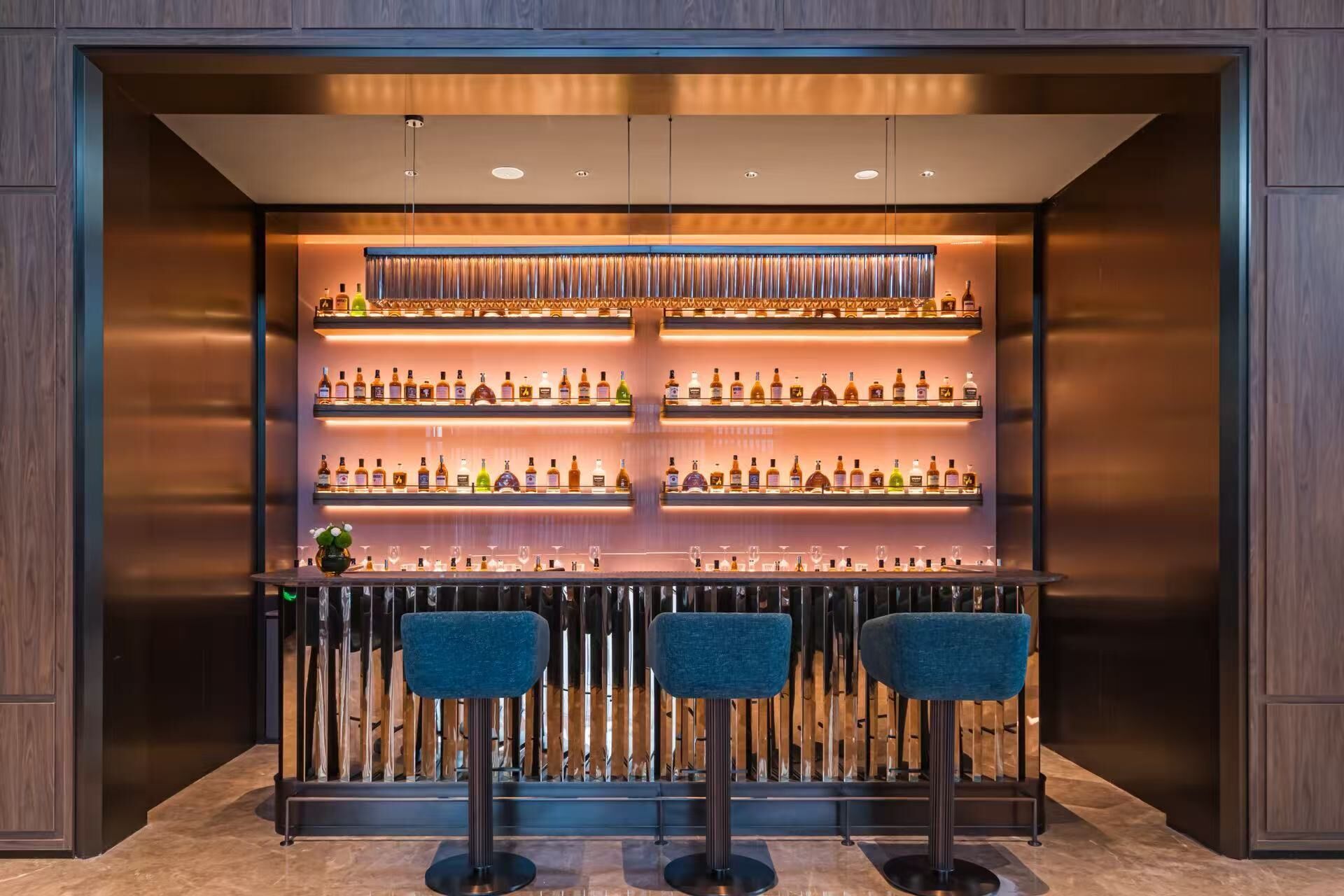 Una zona de bar con varias estanterías llenas de botellas de licor, una barra y tres taburetes azules.
