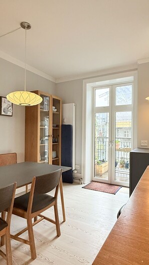 Et spiseområde med et bord og stole, et skab med glaslåger, et hængende lysarmatur og udsigt gennem en glasdør.